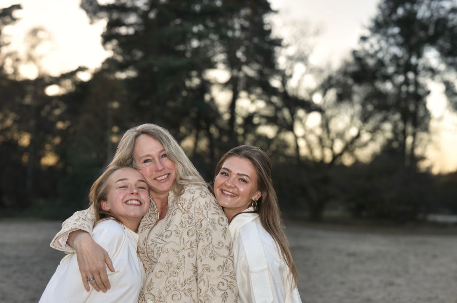 A mother and two daughters smiling and hugging outdoors at sunset in a sandy forest clearing.