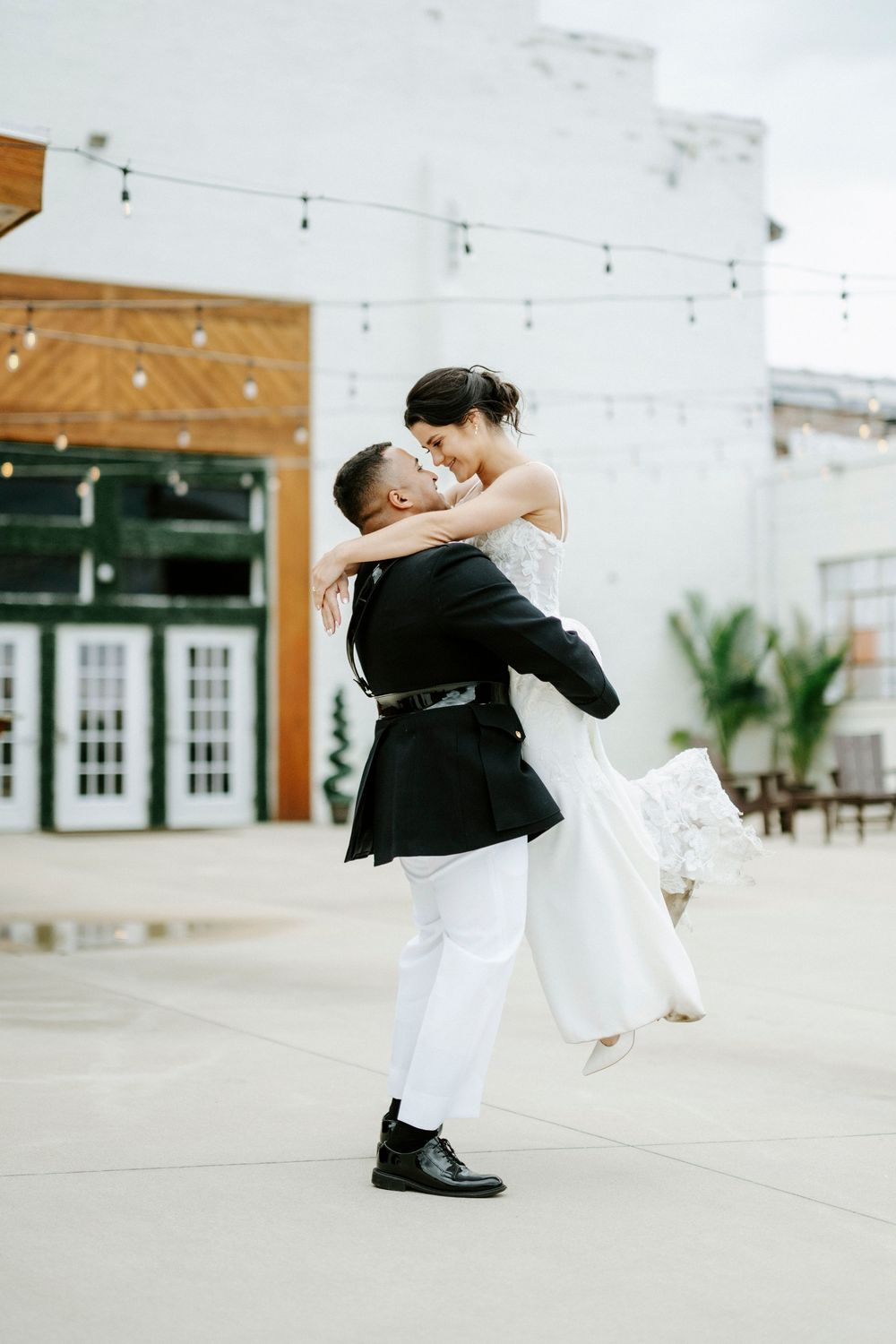 Groom lifts bride in white wedding dress outdoors, sharing a romantic moment in front of a modern venue with string lights.