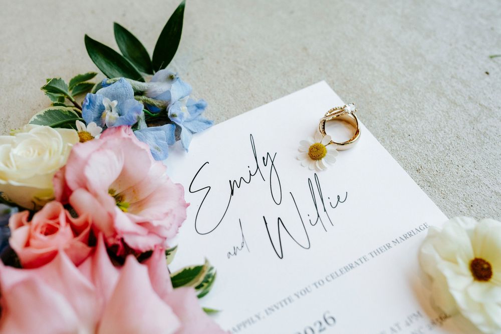 Wedding invitation for Emily and Willie surrounded by pink and blue flowers with gold rings on marble.