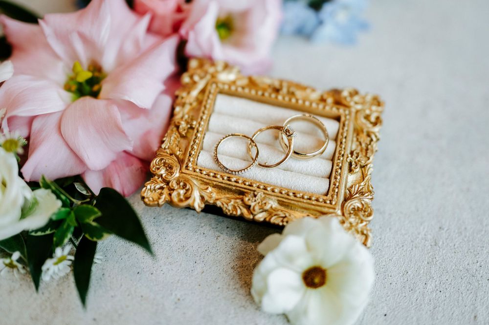 Gold wedding rings displayed in ornate frame surrounded by pink and white flowers.