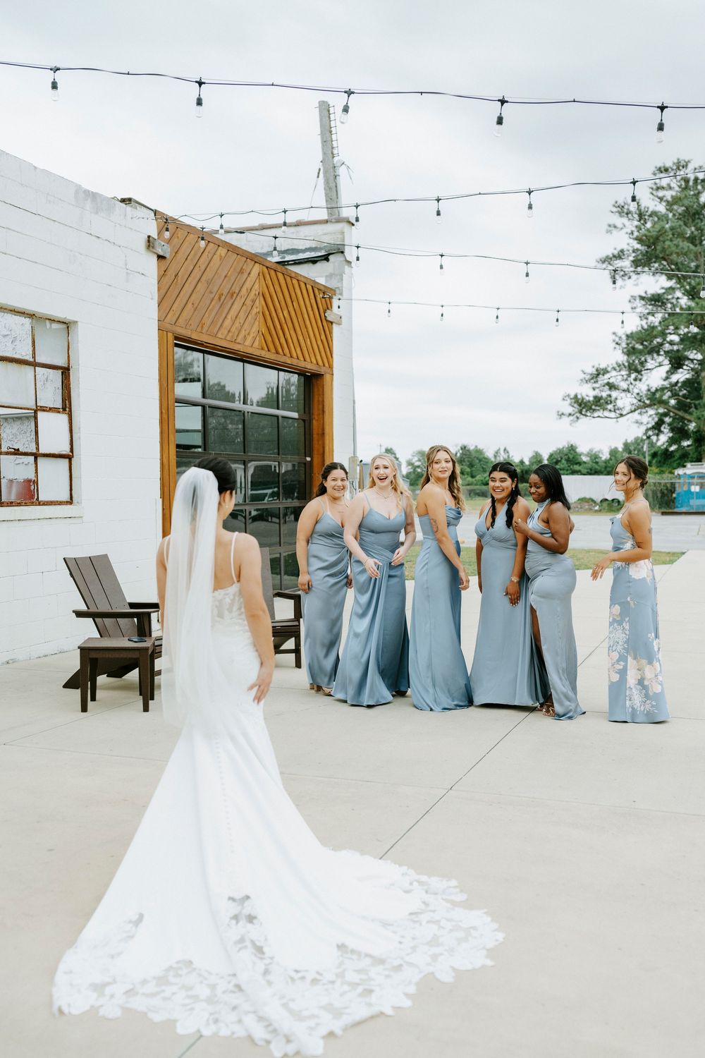 Bride in lace wedding gown with veil faces bridesmaids in dusty blue dresses during outdoor first look.