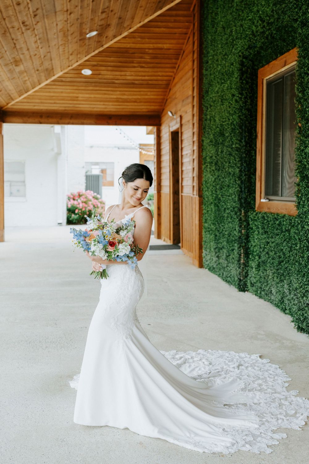 Bride in fitted lace wedding dress holds colorful bouquet beside a green hedge wall at venue.