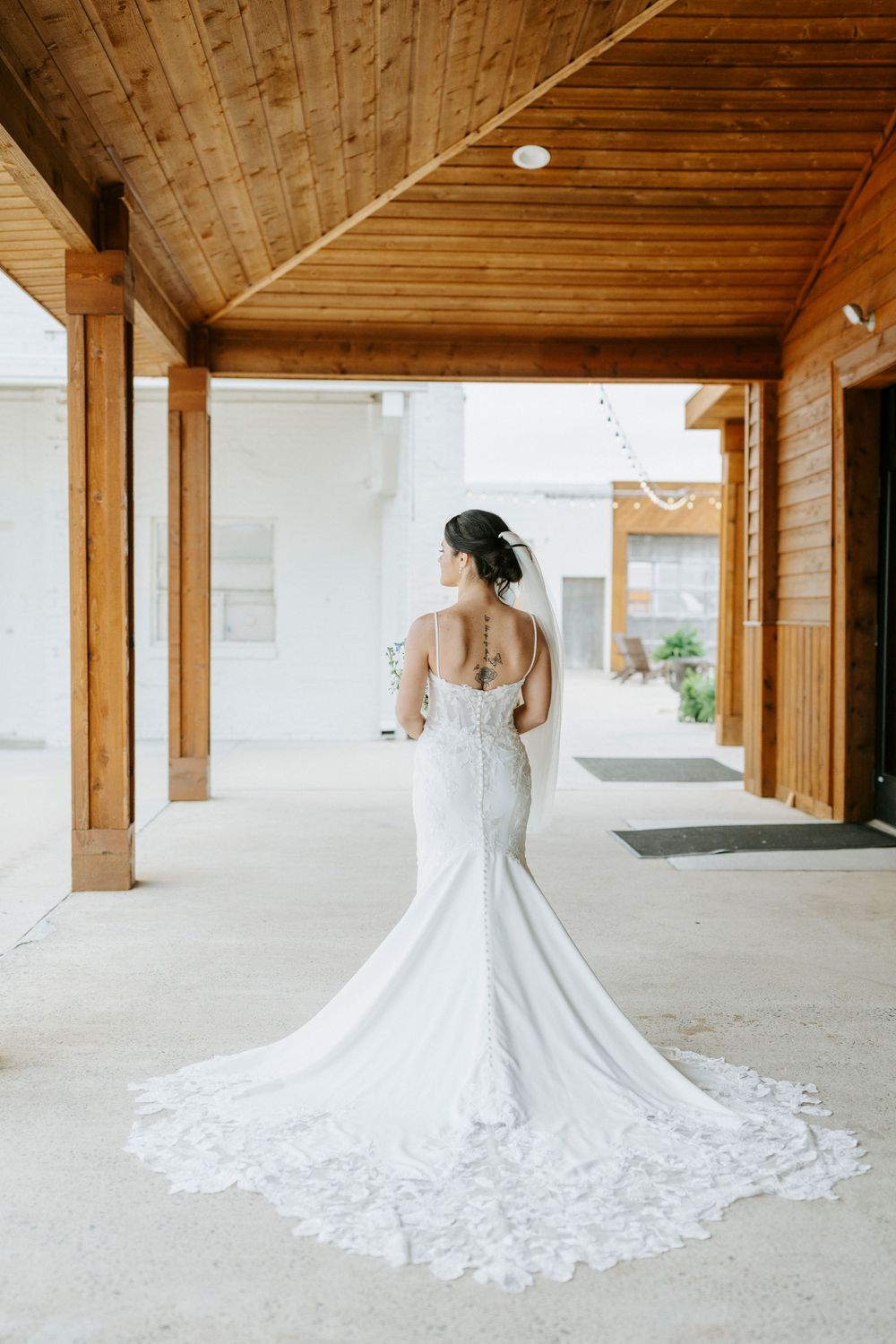 Bride in elegant lace mermaid wedding gown with long train, showing intricate back detail under wooden venue ceiling.