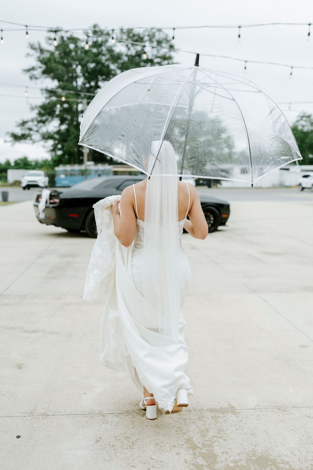 Bride in white gown and veil walking with clear umbrella on rainy wedding day.