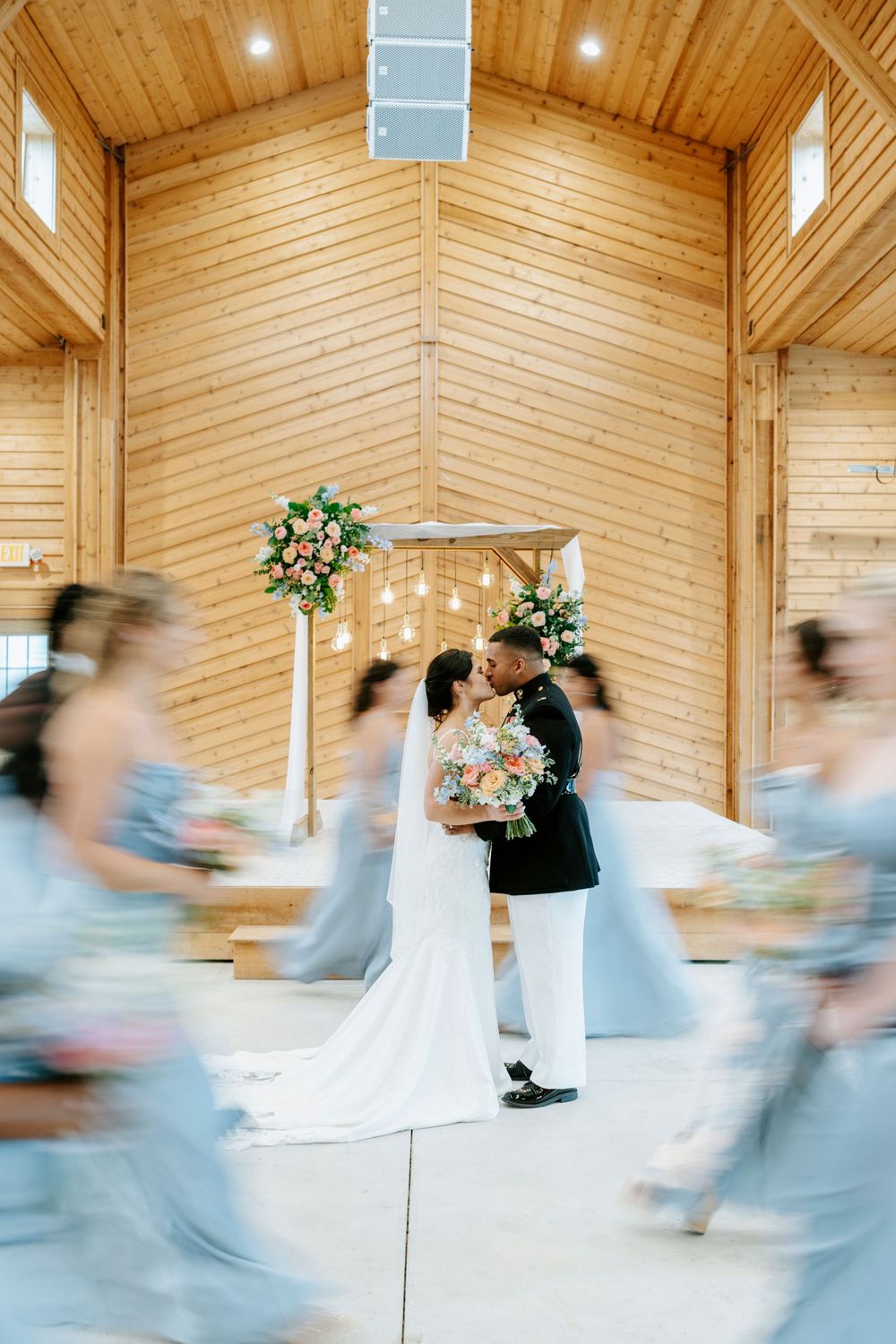 Bride and groom share first kiss in rustic wood chapel, surrounded by bridesmaids in flowing blue dresses with floral arch backdrop.