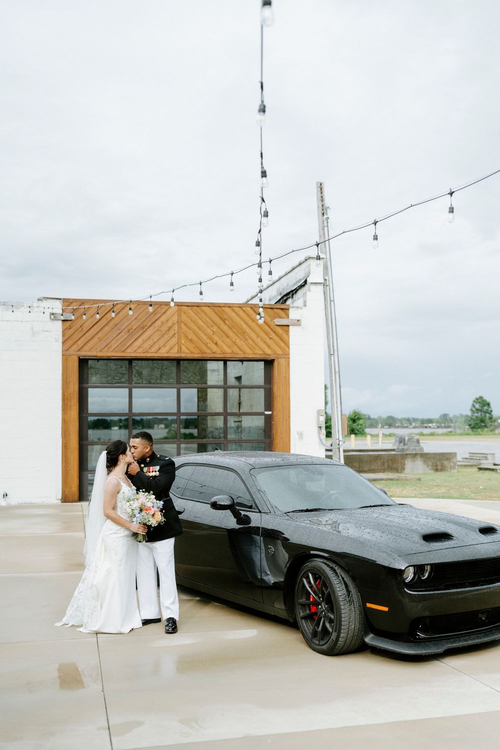 Bride and groom pose beside a black Dodge Challenger at an outdoor wedding venue with string lights overhead.