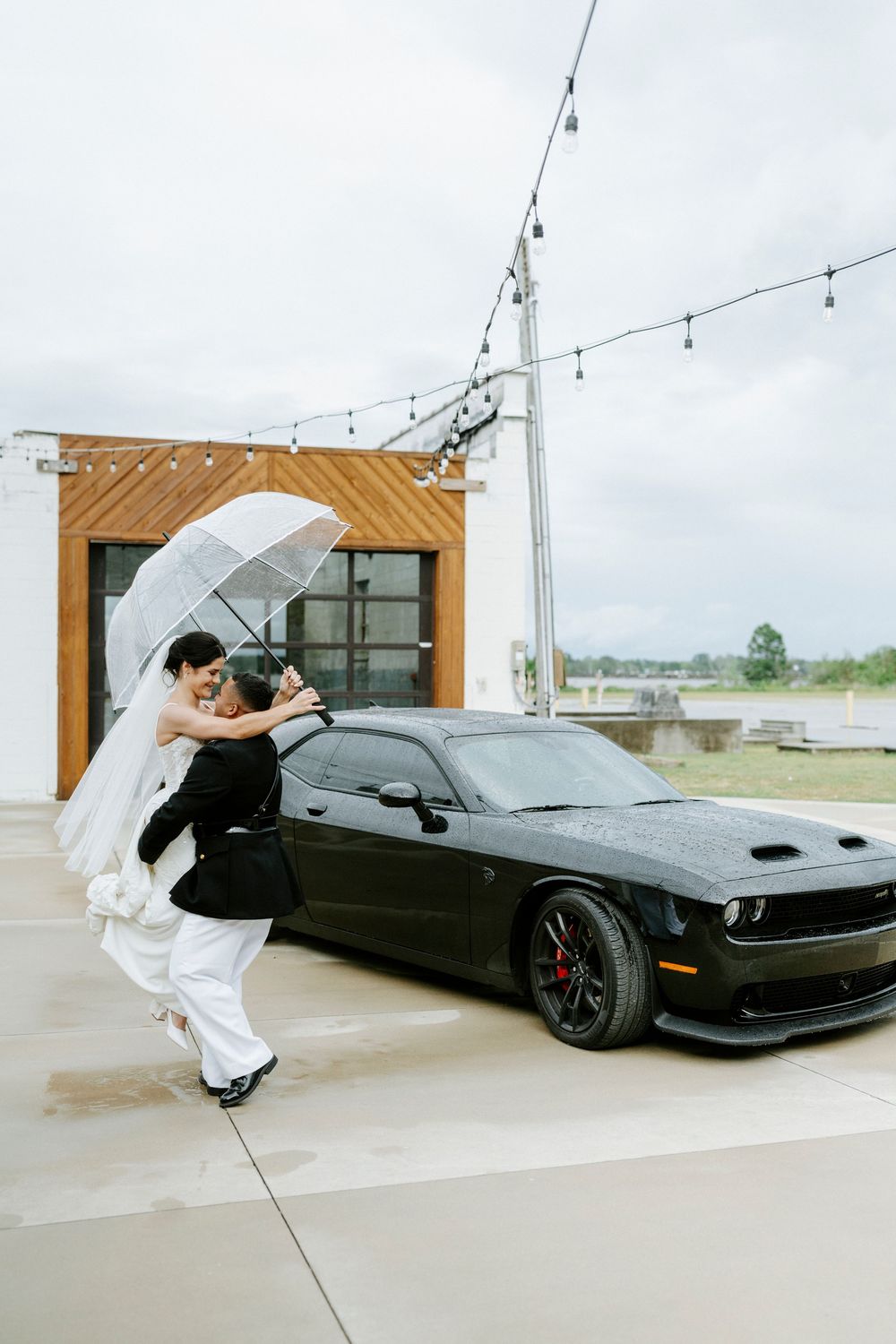 Bride and groom embrace under a clear umbrella beside a black Dodge Challenger on their rainy wedding day.