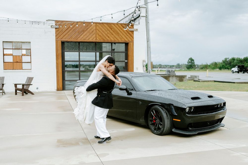 Bride and groom sharing a romantic dip kiss in front of a black Dodge Challenger muscle car at their wedding venue.