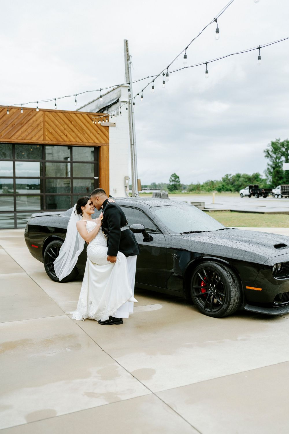 Bride and groom sharing a romantic kiss beside a black Dodge Challenger at an outdoor wedding venue.