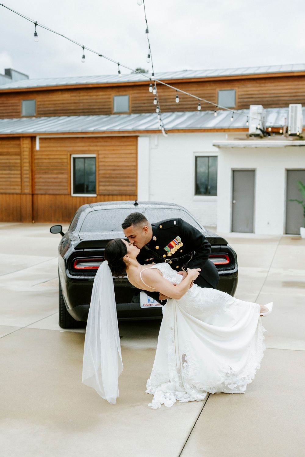 Military groom dips bride in white mermaid gown for romantic kiss beside black car at rustic wedding venue.