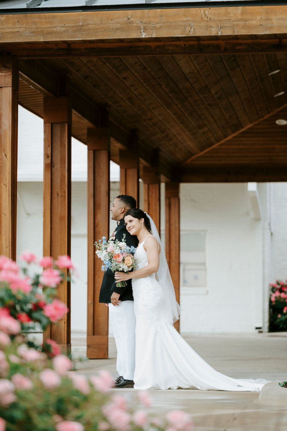 Bride in fitted white gown with veil holding bouquet stands with groom in military dress uniform among pink roses.