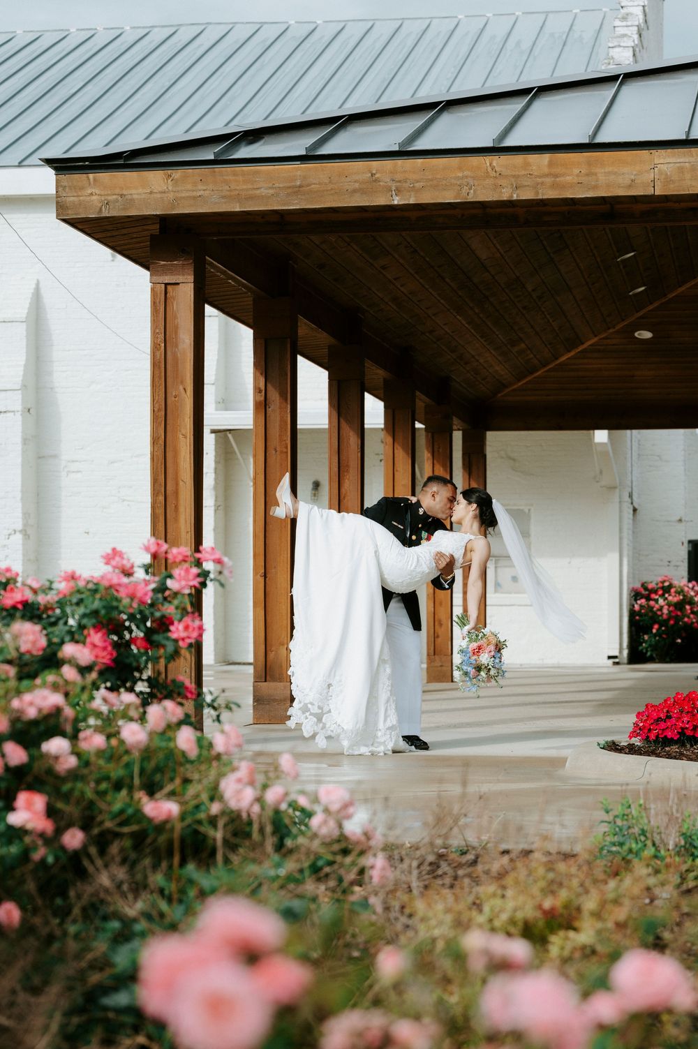 Groom in military uniform dips bride in lace gown for a romantic kiss amid blooming roses.