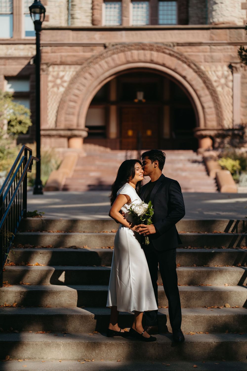 Couple in elegant wedding attire sharing a romantic moment on stone steps before a historic brick building.