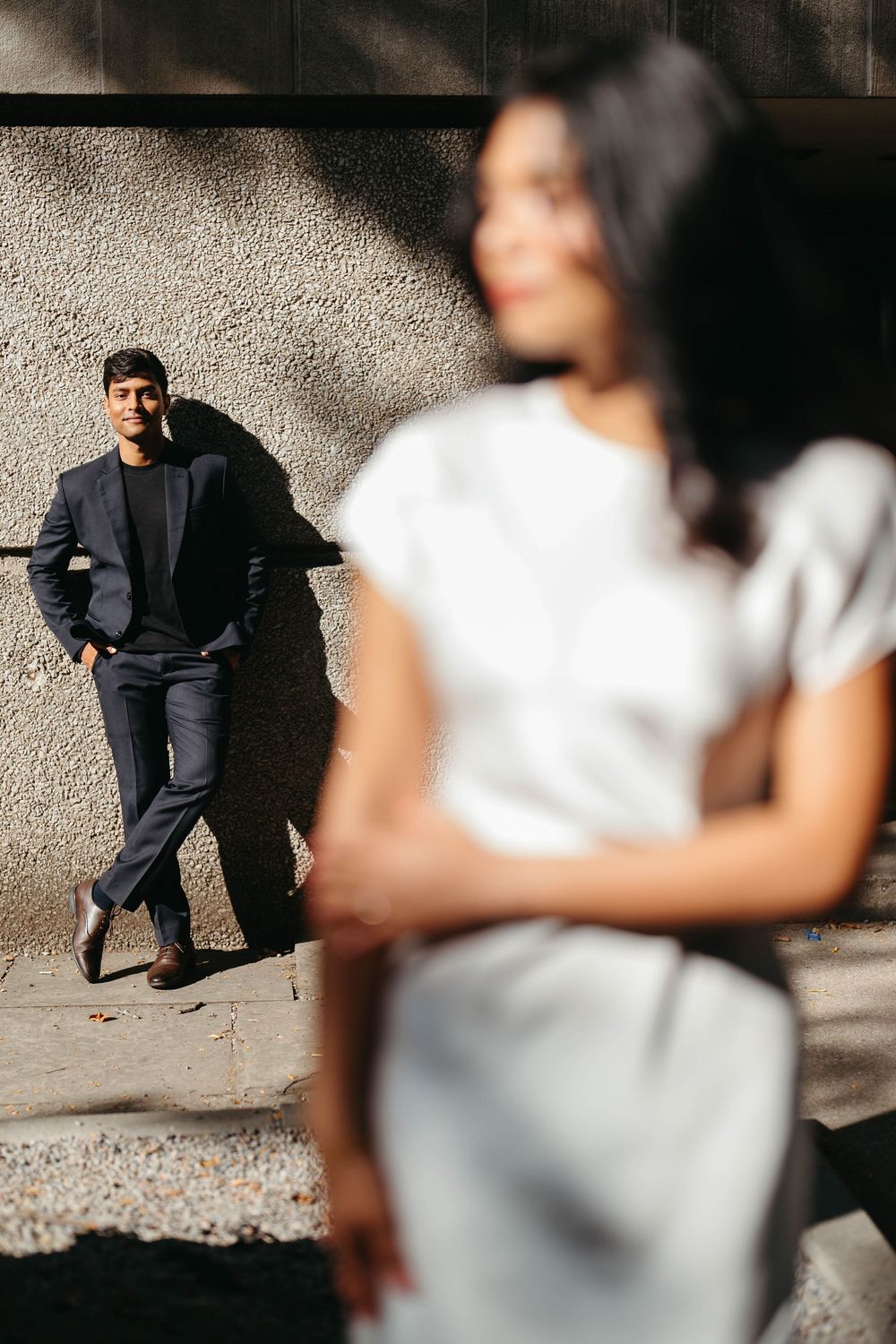 A man in a navy suit leans against a textured wall while a woman in a white dress stands blurred in the foreground.
