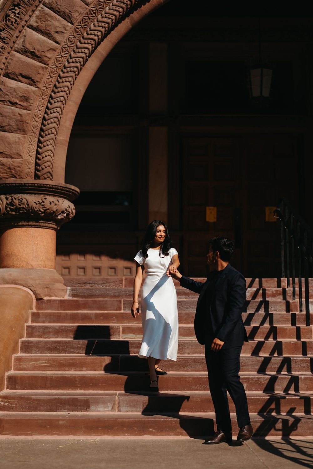 Couple in elegant attire on historic stone steps under ornate arch, woman in white dress holding man's hand in navy suit.
