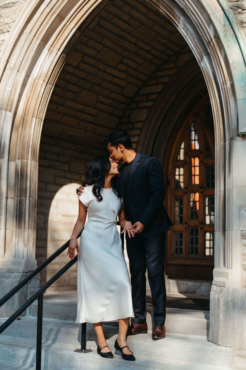 Couple sharing a romantic kiss under a stone Gothic arch doorway, woman in white dress, man in dark suit.