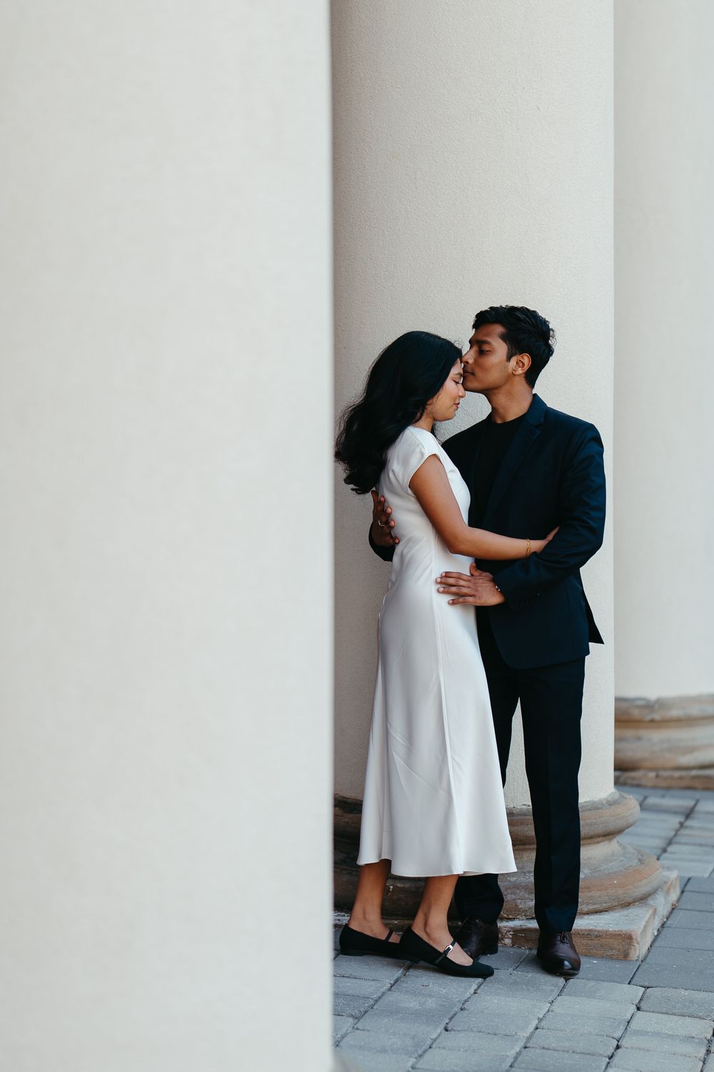 Couple sharing a romantic kiss between white columns, woman in white midi dress, man in dark suit.