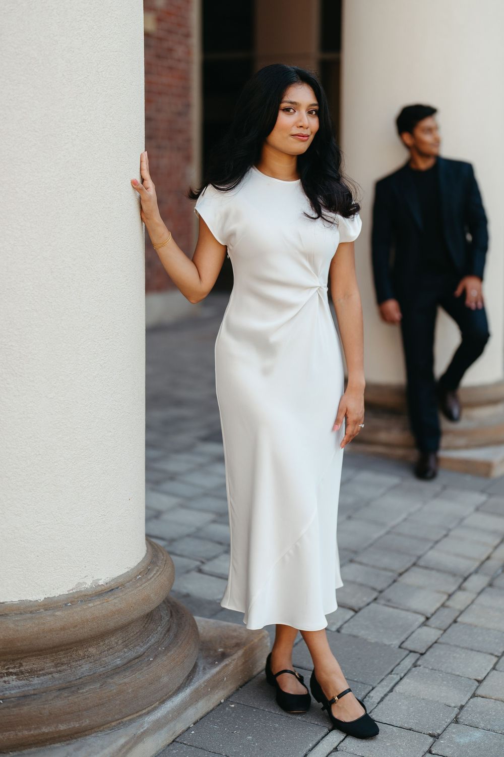Elegant woman in white satin midi dress posing by column, man in navy suit walking in background on cobblestone.