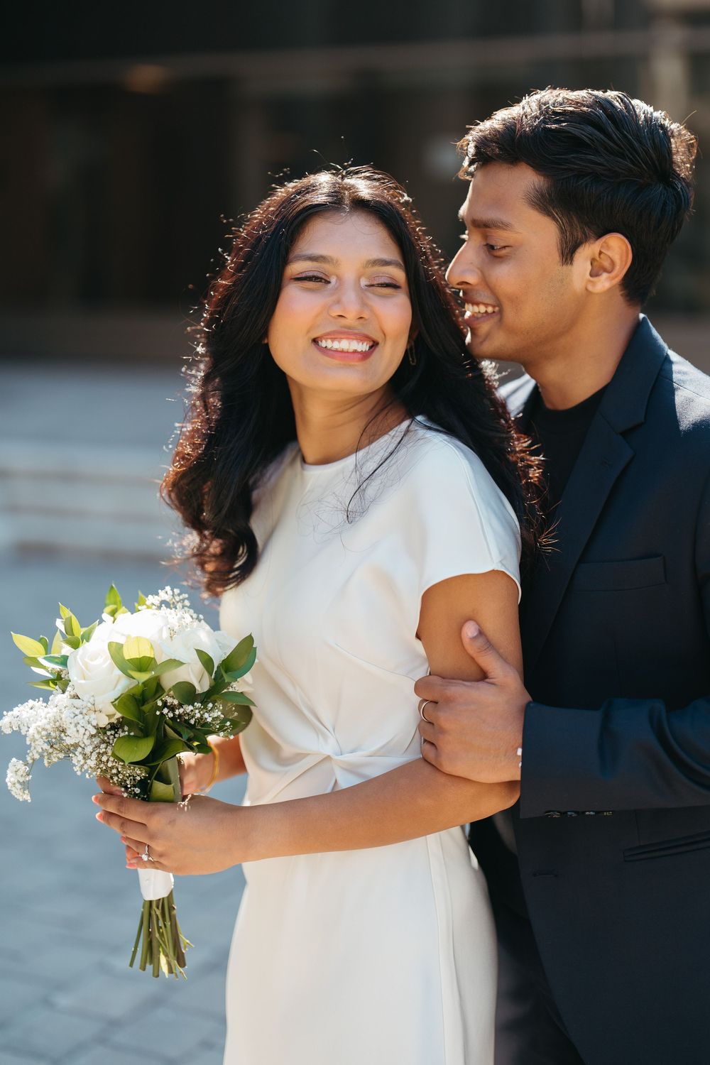 Happy bride in white dress holds bouquet while groom in dark suit embraces her from behind outdoors.