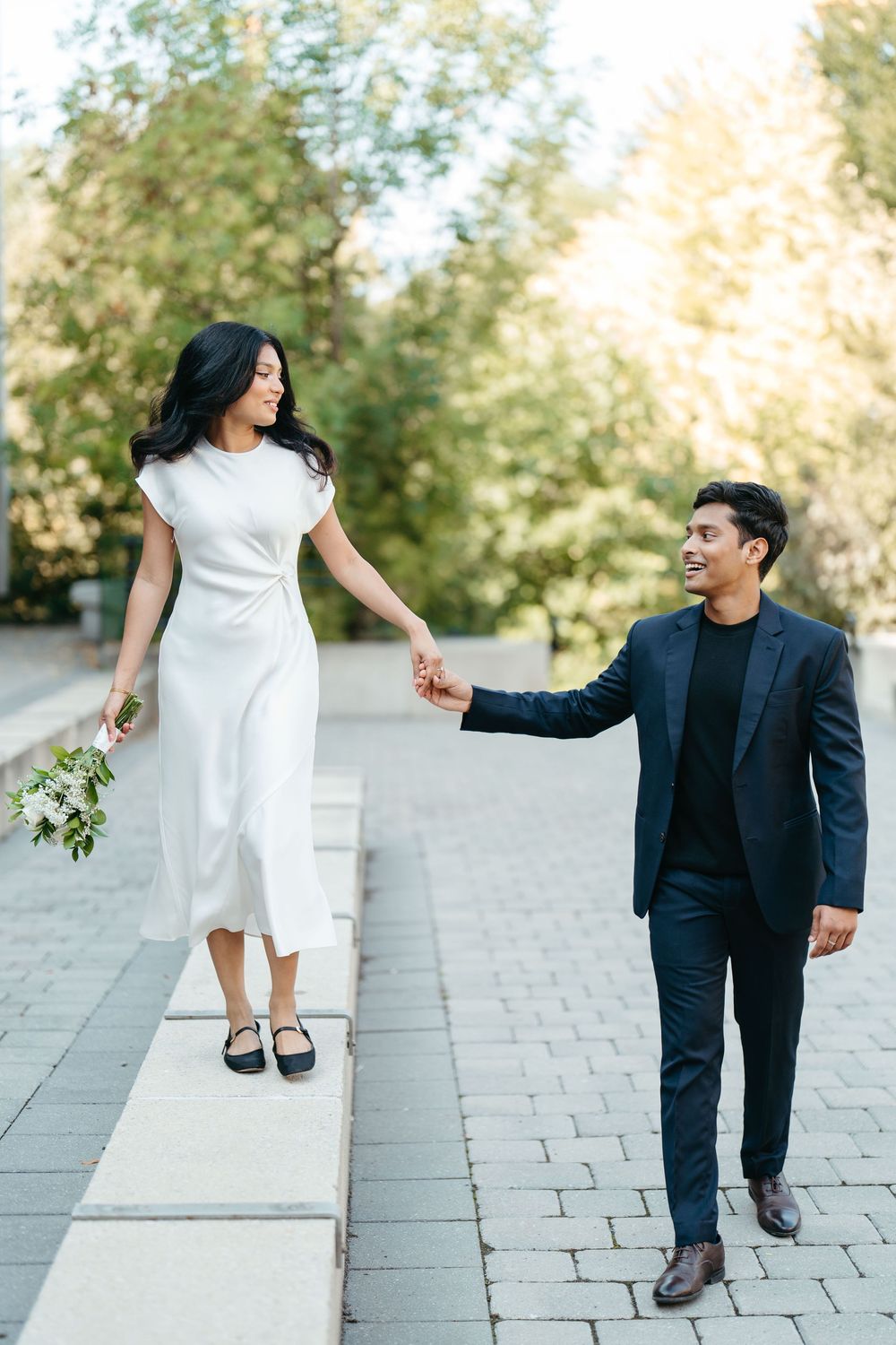Couple in wedding attire holding hands while walking outdoors, bride in white dress holding bouquet, groom in navy suit.