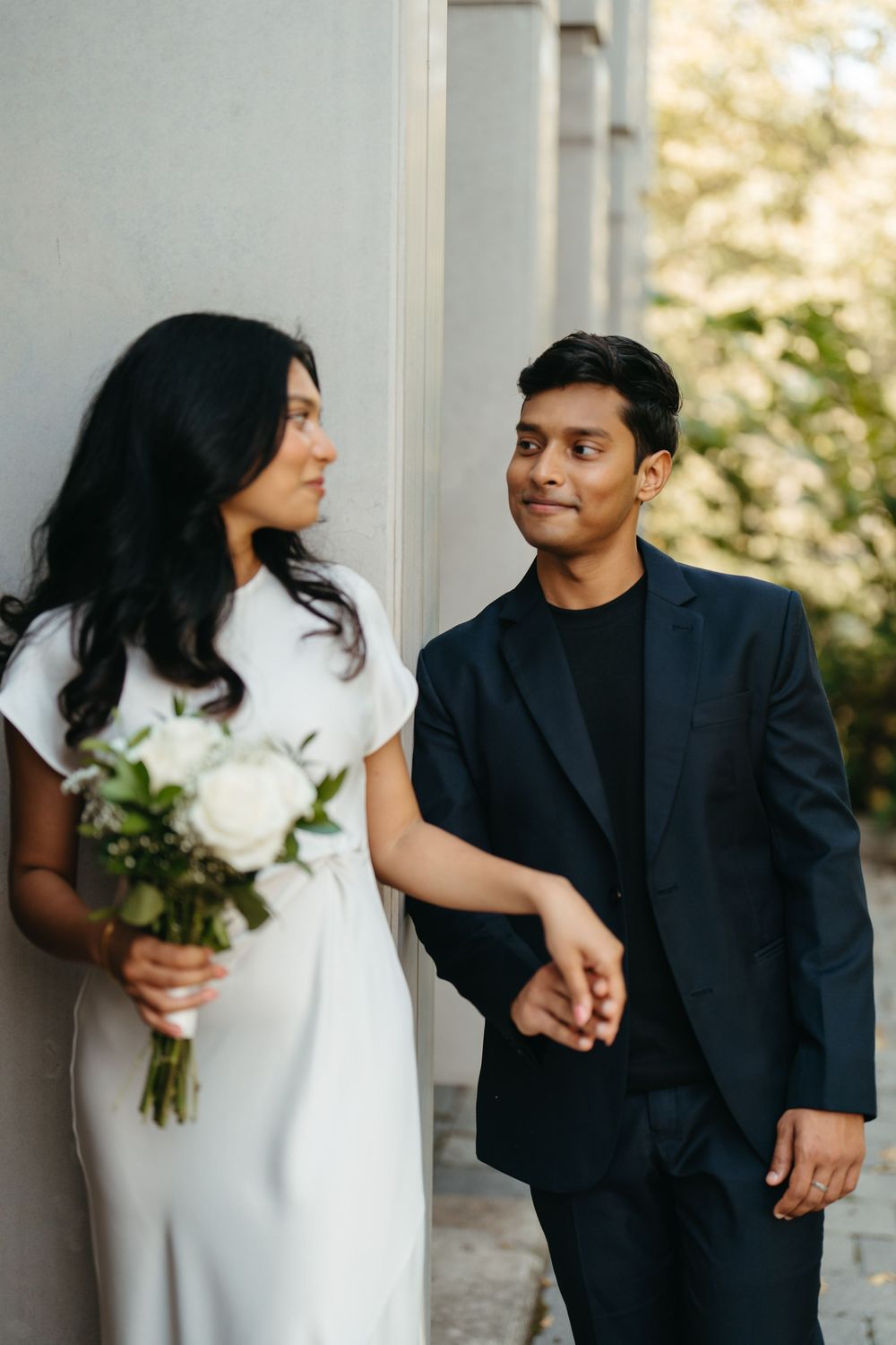 Couple in wedding attire holding hands, bride in white dress with bouquet, groom in dark suit, outdoors.
