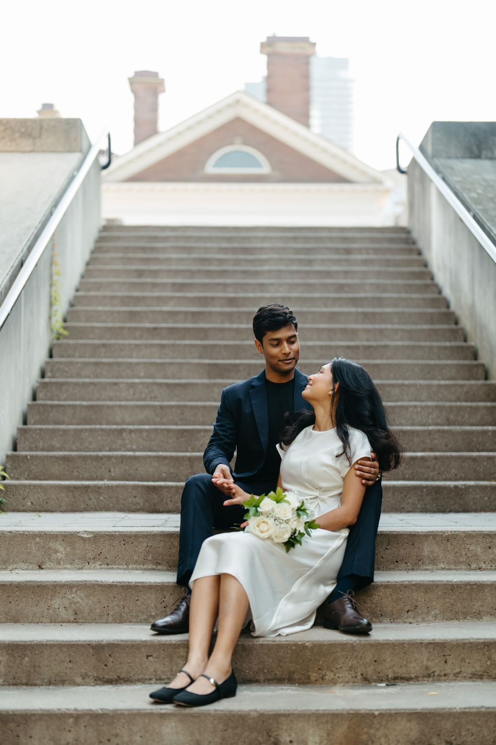 Couple in wedding attire sitting on outdoor steps, bride in white dress holding bouquet, groom in navy suit.