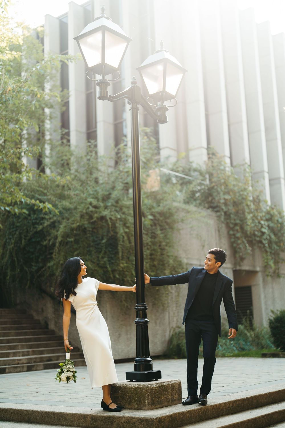 Bride in white dress and groom in dark suit posing by a street lamp in an urban outdoor setting.