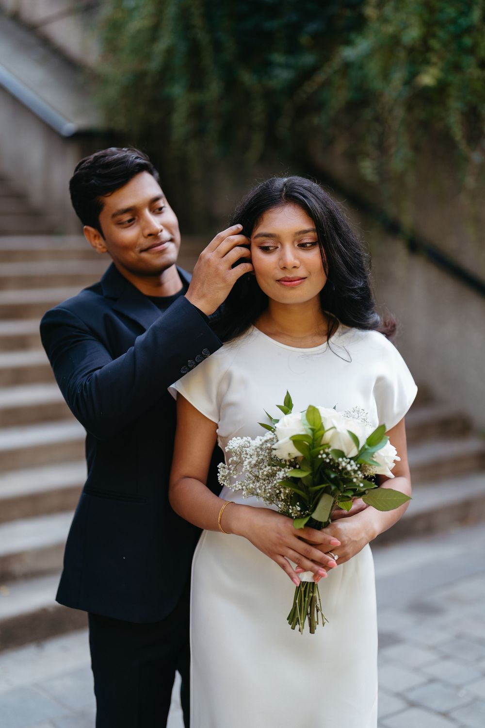 A groom in a black suit adjusts his bride's hair as she holds a white floral bouquet in an outdoor setting.