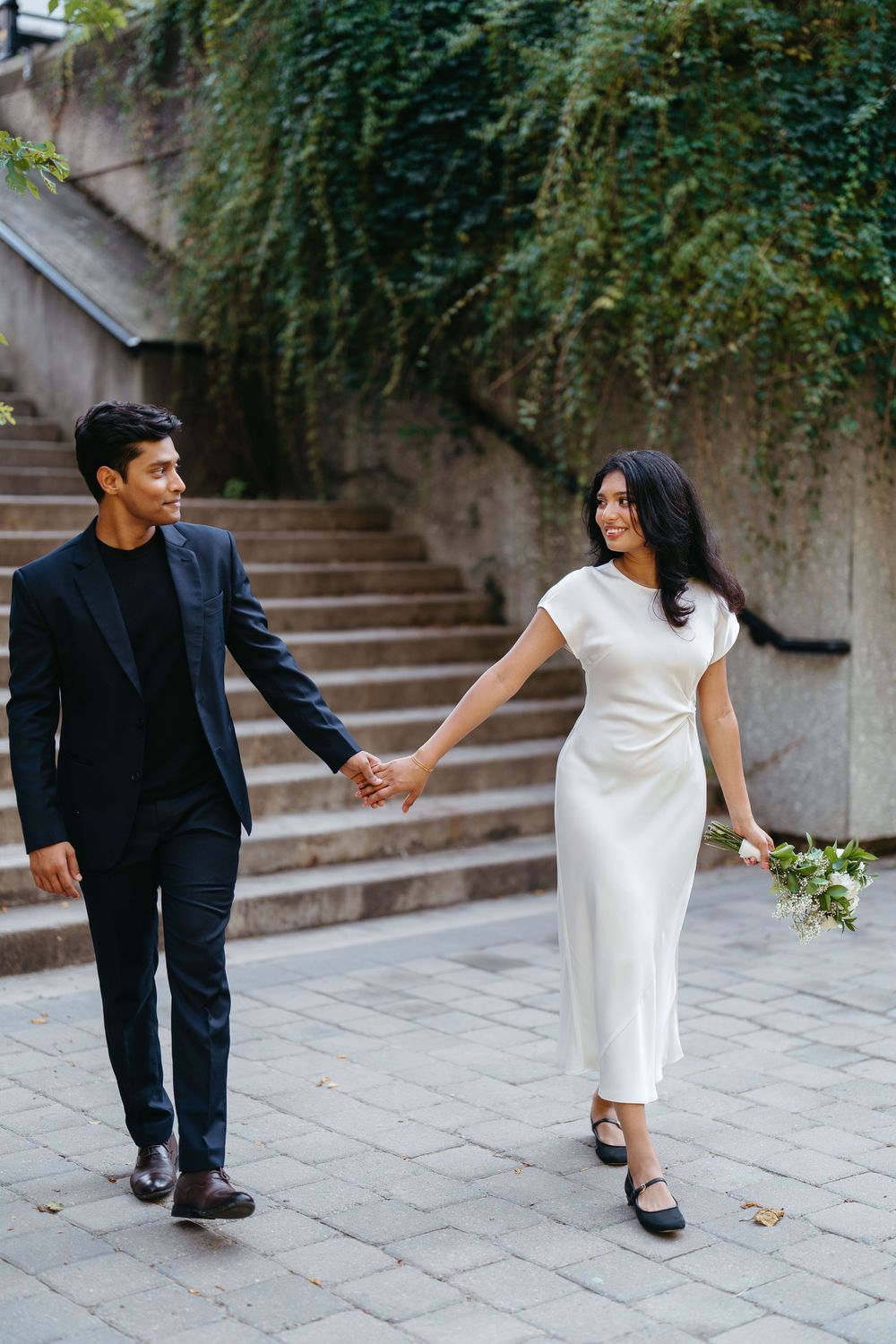 Couple in elegant attire holding hands outdoors, woman in white dress with bouquet, man in black suit near stone steps.