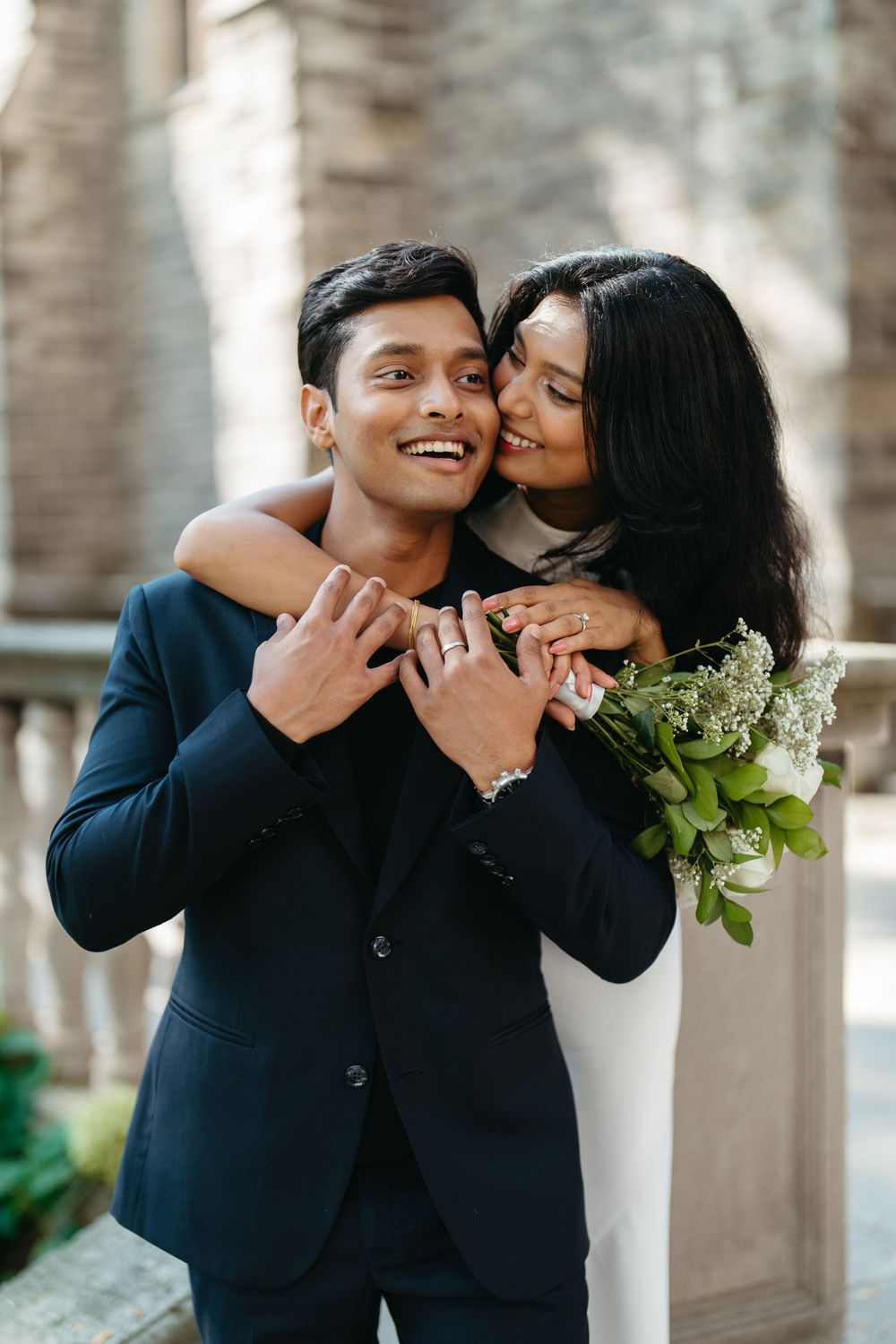 Smiling groom in navy suit embraced by bride in white dress holding bouquet outside historic stone building.