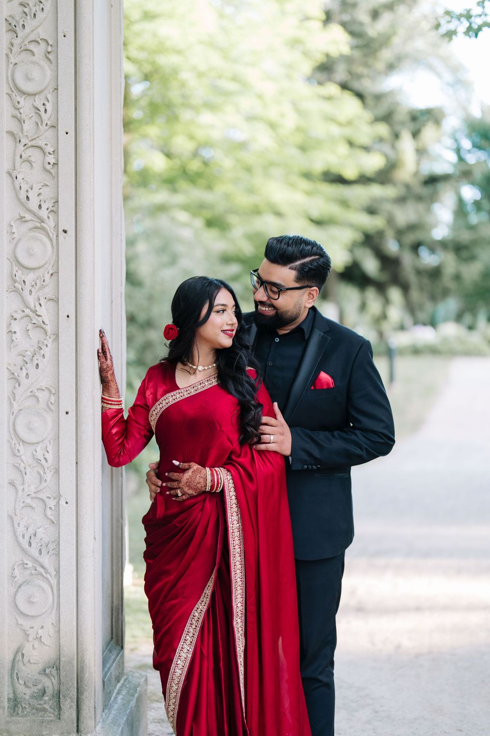 Couple in traditional Indian attire, woman in red saree and man in black suit, posing near ornate white column outdoors.
