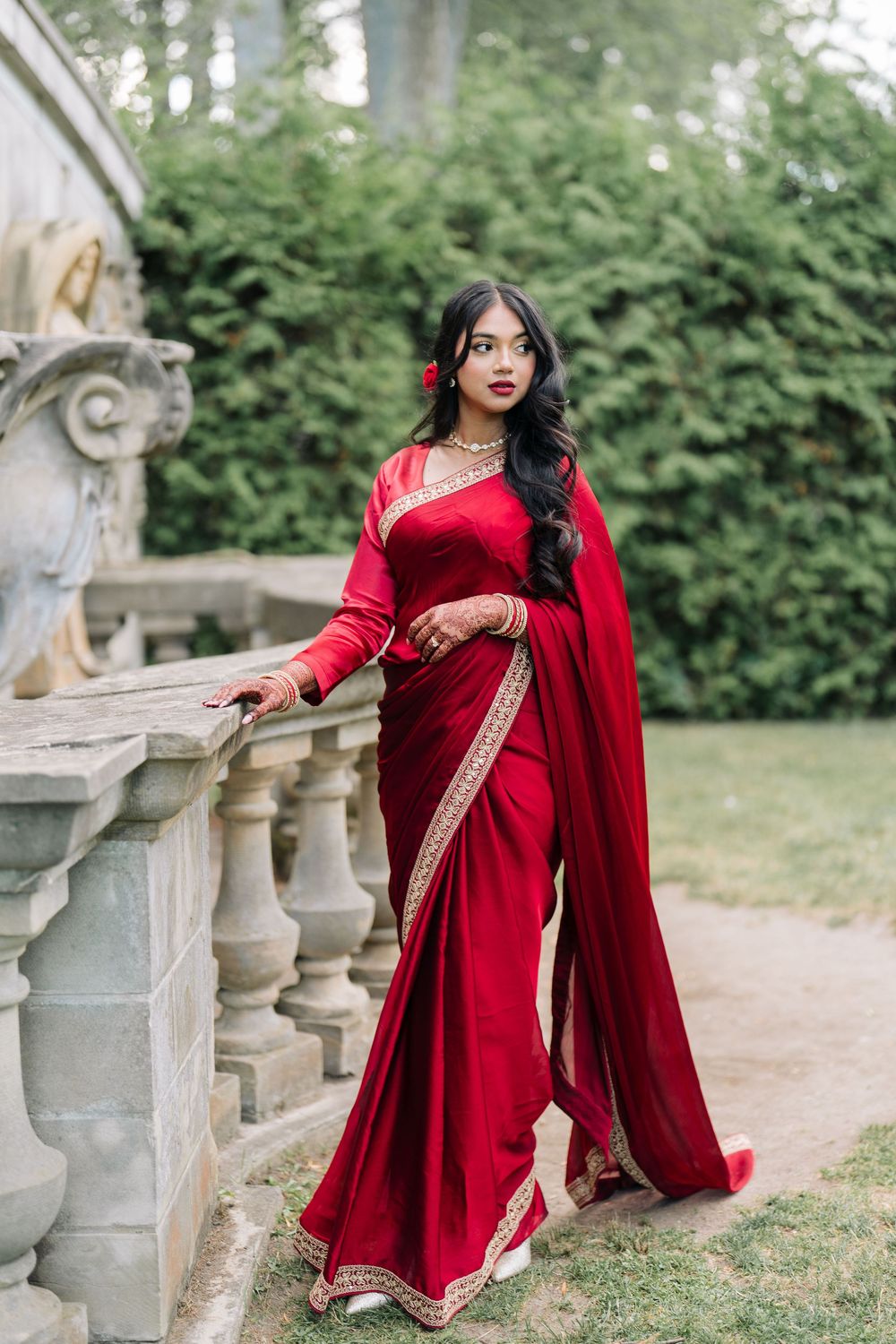 Woman in elegant red saree with gold trim poses gracefully by stone balustrade in lush garden setting.