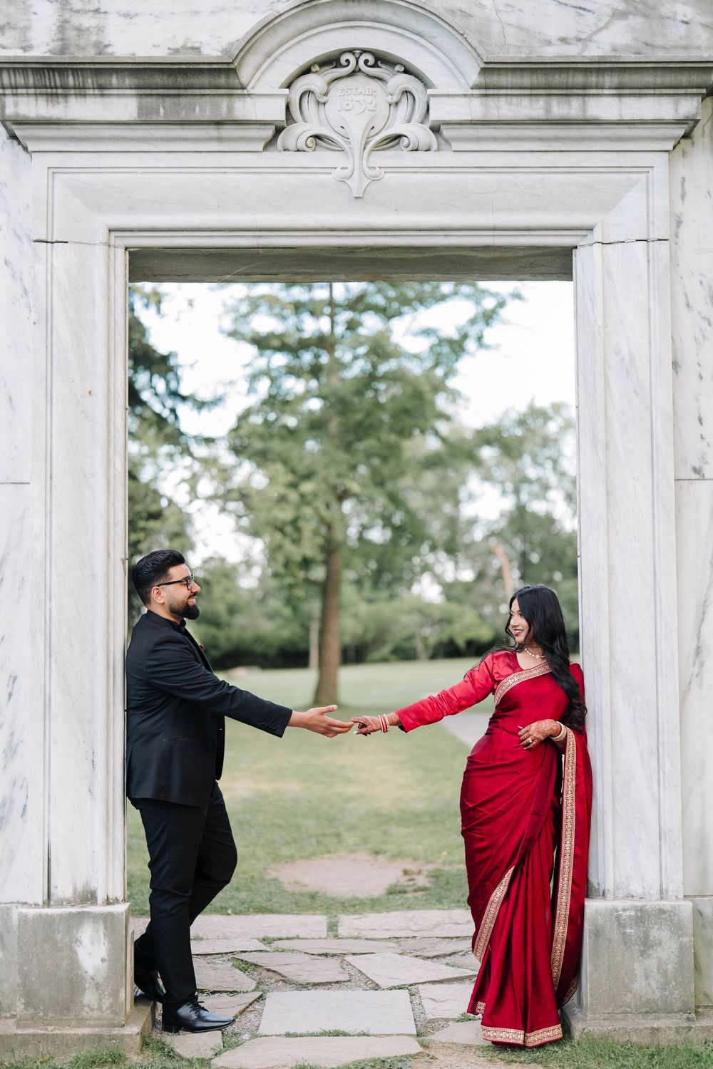 A couple in traditional Indian attire holds hands under an ornate stone archway in a lush garden setting.