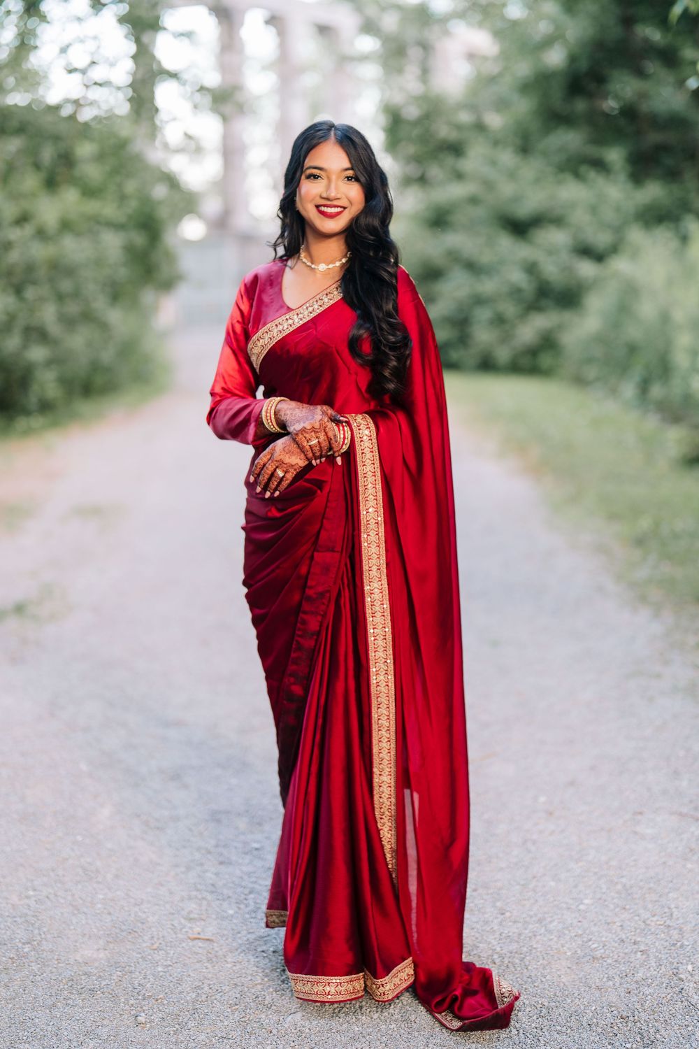 Woman in elegant red saree with gold border and traditional jewelry poses on a garden path.