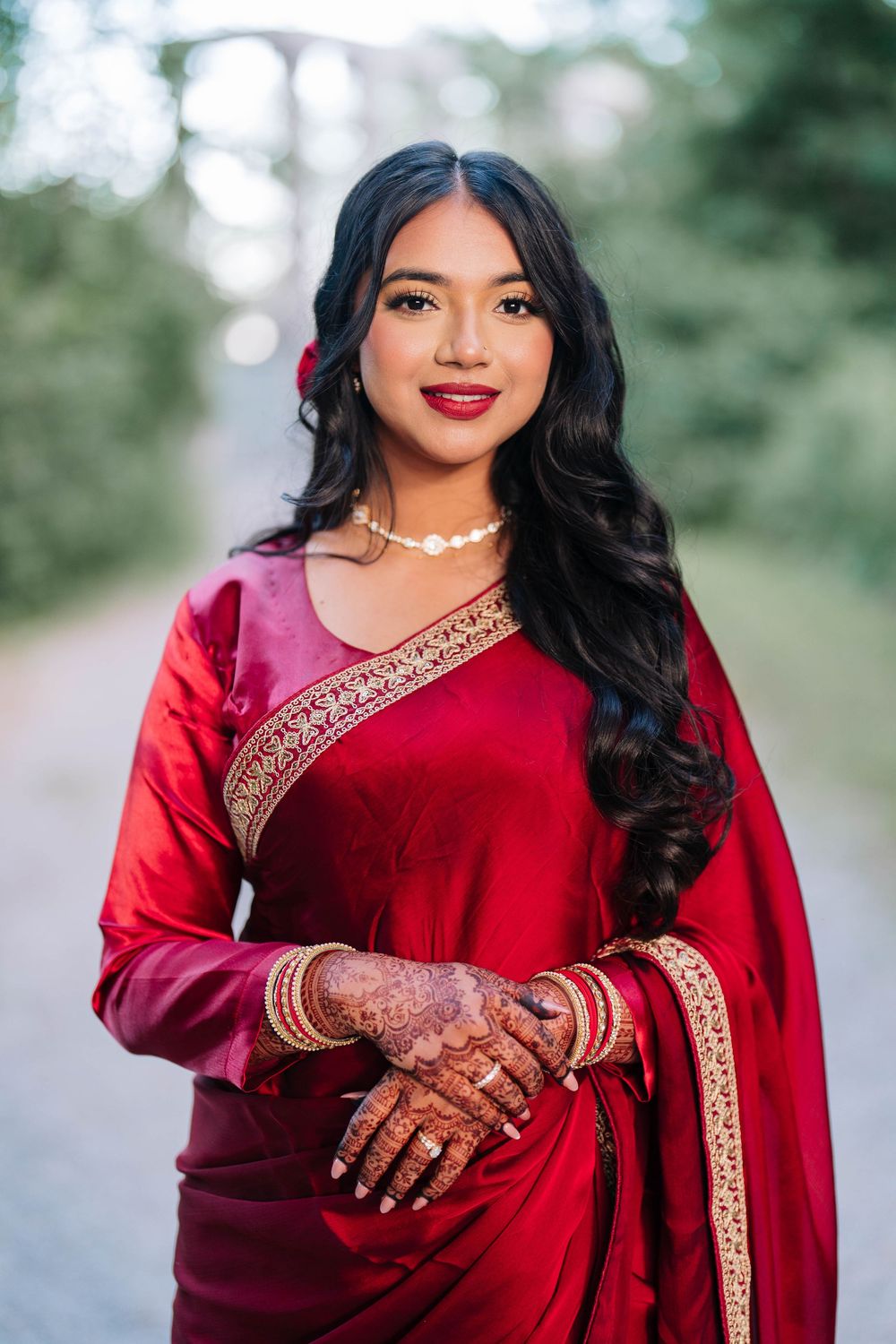 Young woman in red saree with gold trim, henna hands, pearl necklace, and red flower in dark wavy hair, smiling outdoors.