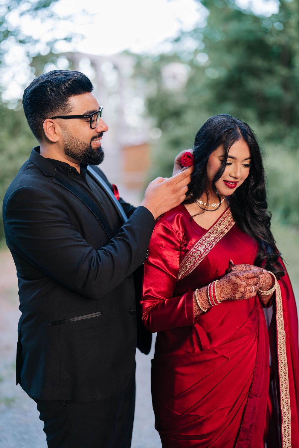 Indian couple in traditional wedding attire, man in black suit adjusting womans hair, bride in red saree with mehndi.