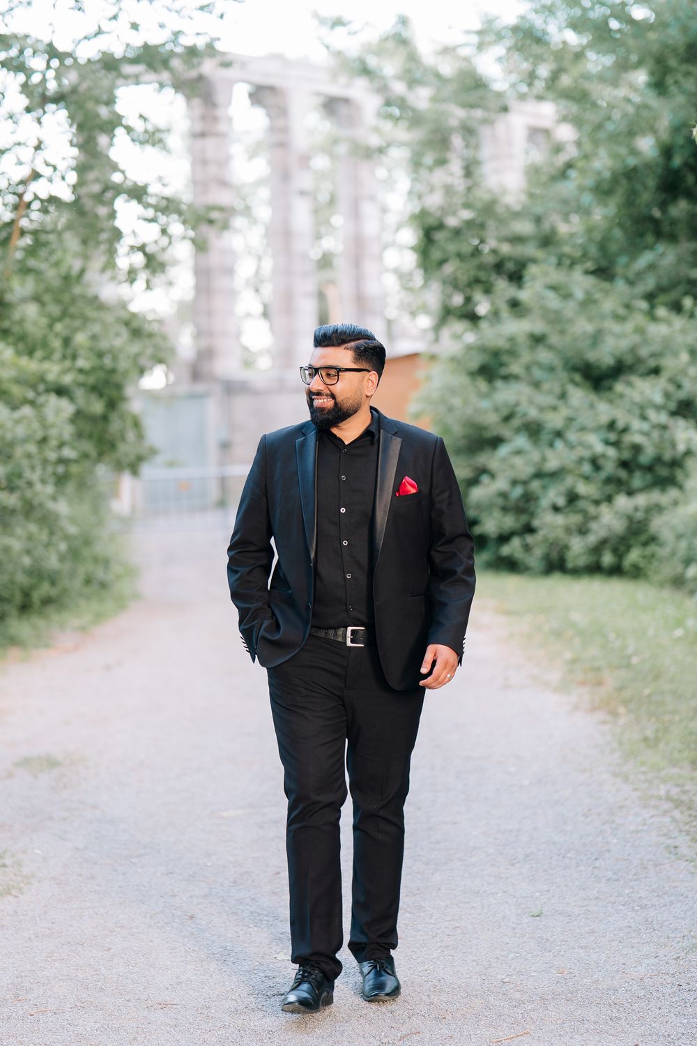 A well-dressed man in a black suit with red pocket square walks confidently along a tree-lined path.