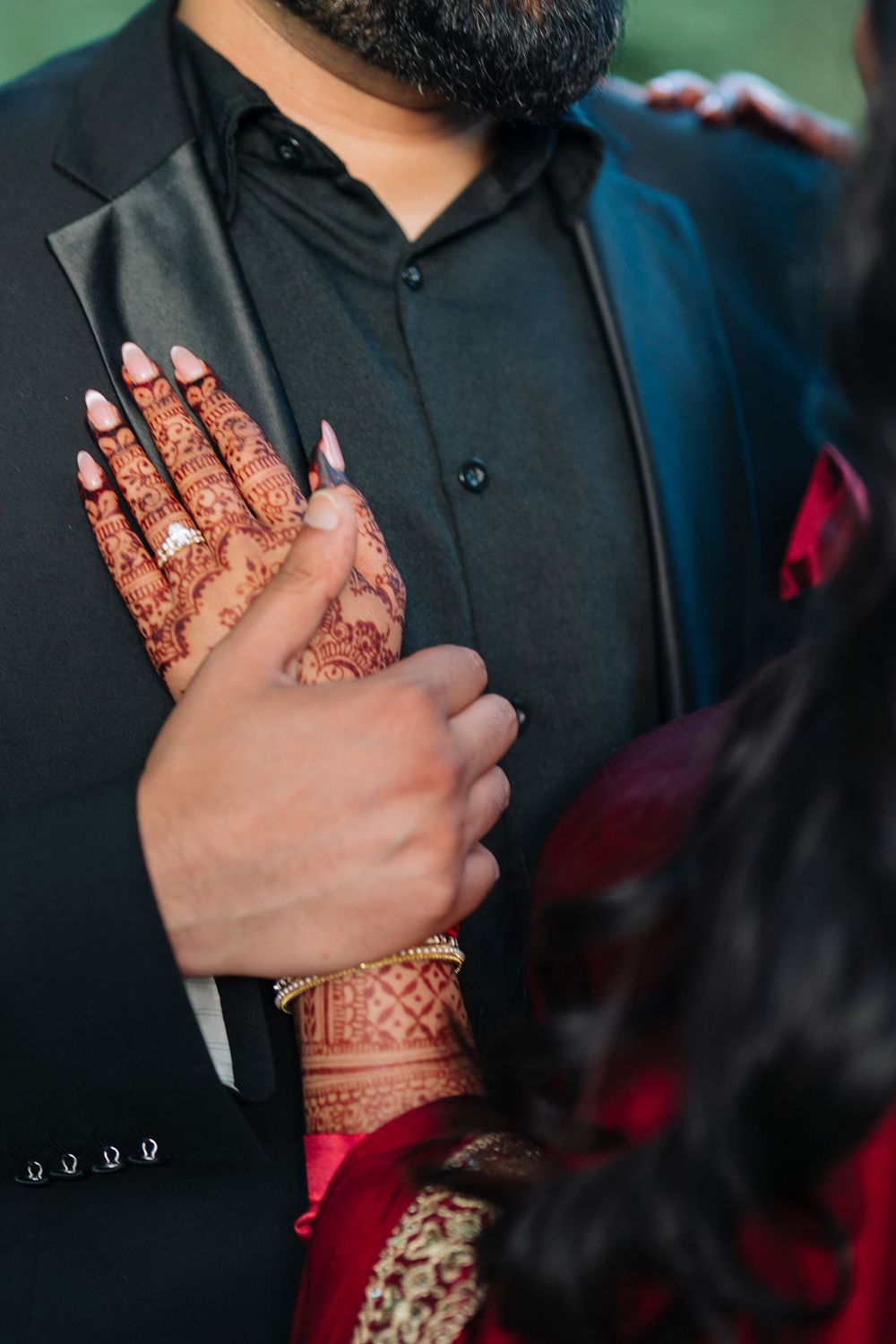 Bride with intricate henna mehndi designs and ring rests hand on groom's black sherwani suit at Indian wedding.