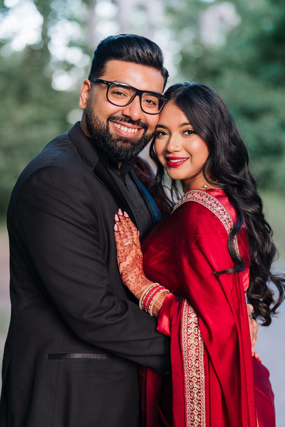 Smiling Indian couple in traditional wedding attire, man in black suit and woman in red saree with mehndi, posing outdoors.