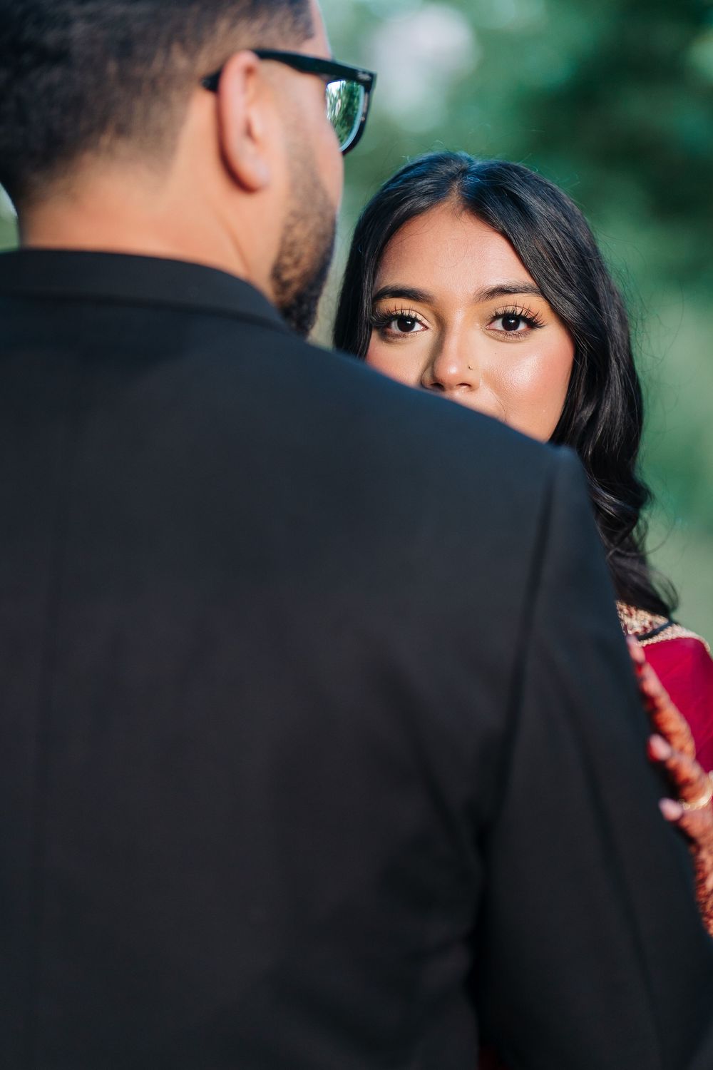 Indian bride in red saree gazing at groom in black suit during outdoor wedding portrait session.