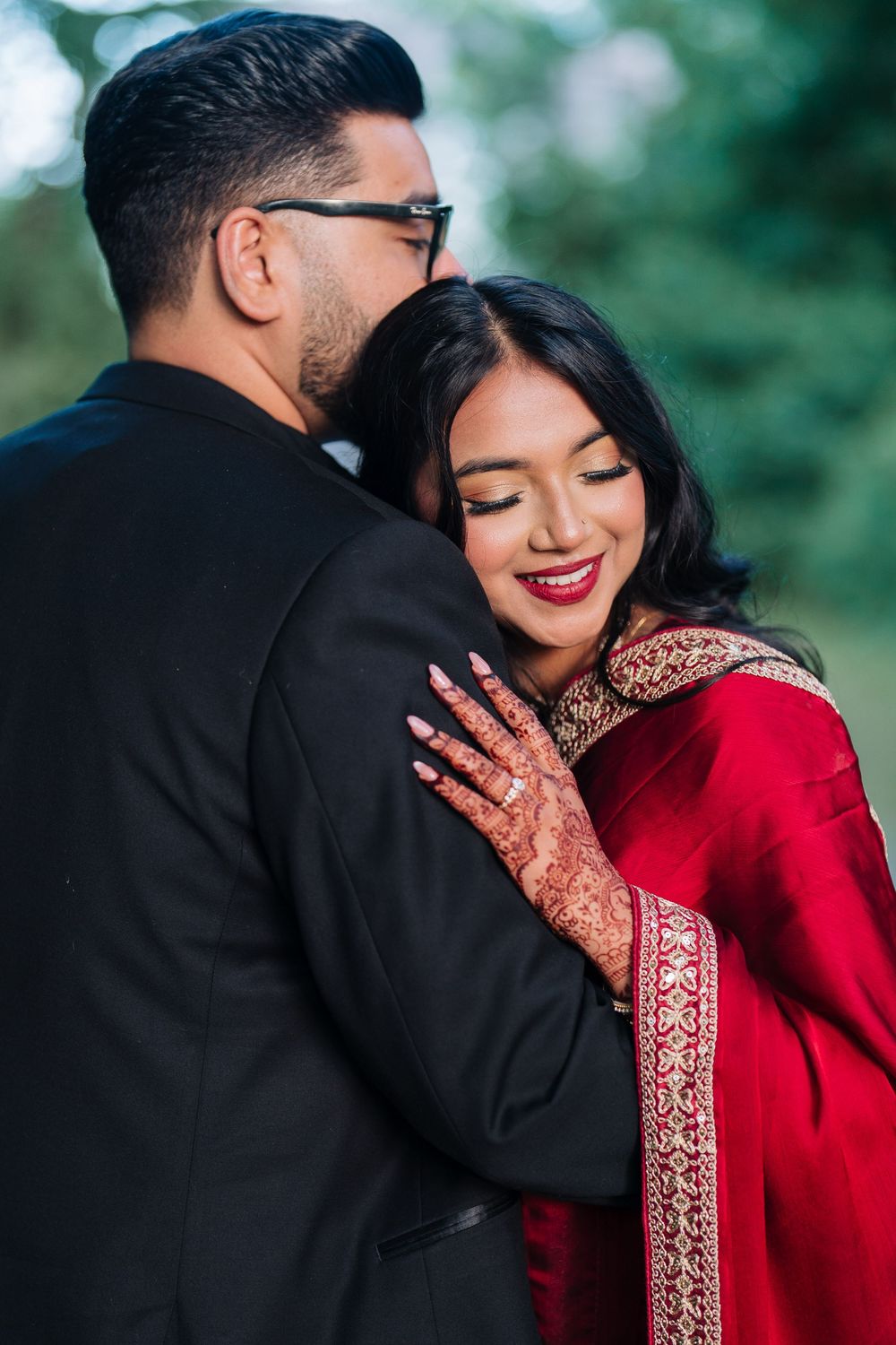 Couple in traditional Indian wedding attire, bride in red saree with henna hands embracing groom in black suit outdoors.