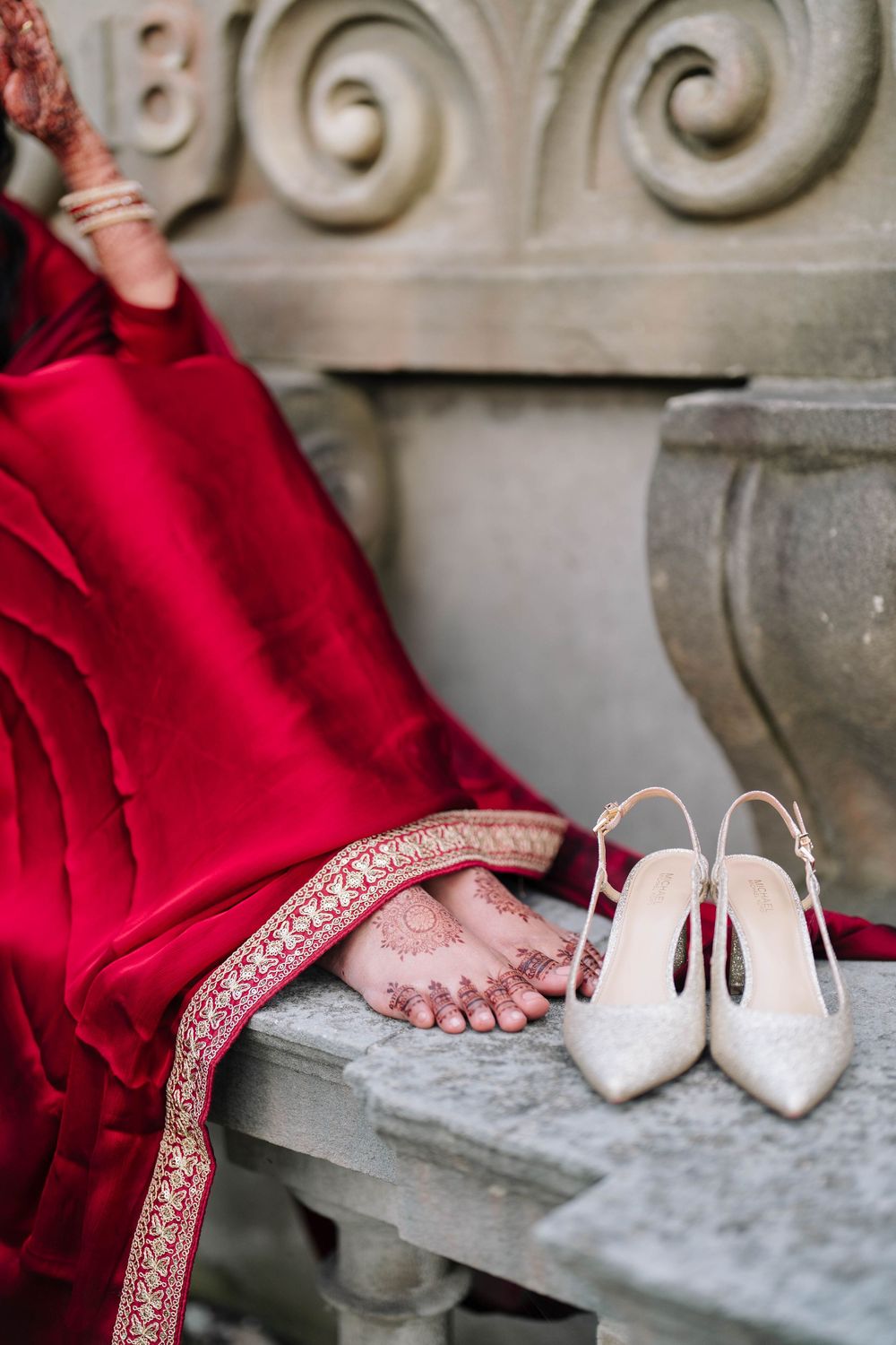 Bride in red lehenga with mehndi on feet beside white slingback heels on stone steps.