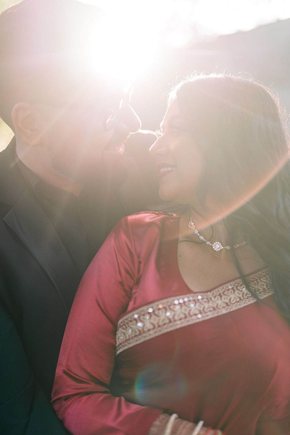 Couple in romantic embrace with sun flare, woman in red traditional dress with silver embroidery, man in dark suit.