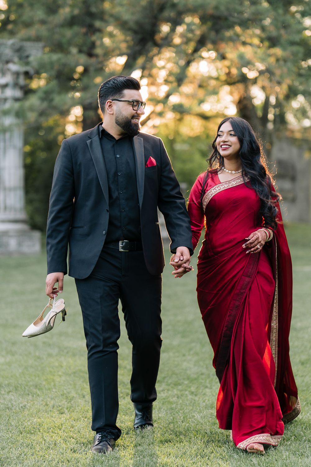 Couple in formal attire walking outdoors, man in black suit holding heels, woman in red saree, smiling together.