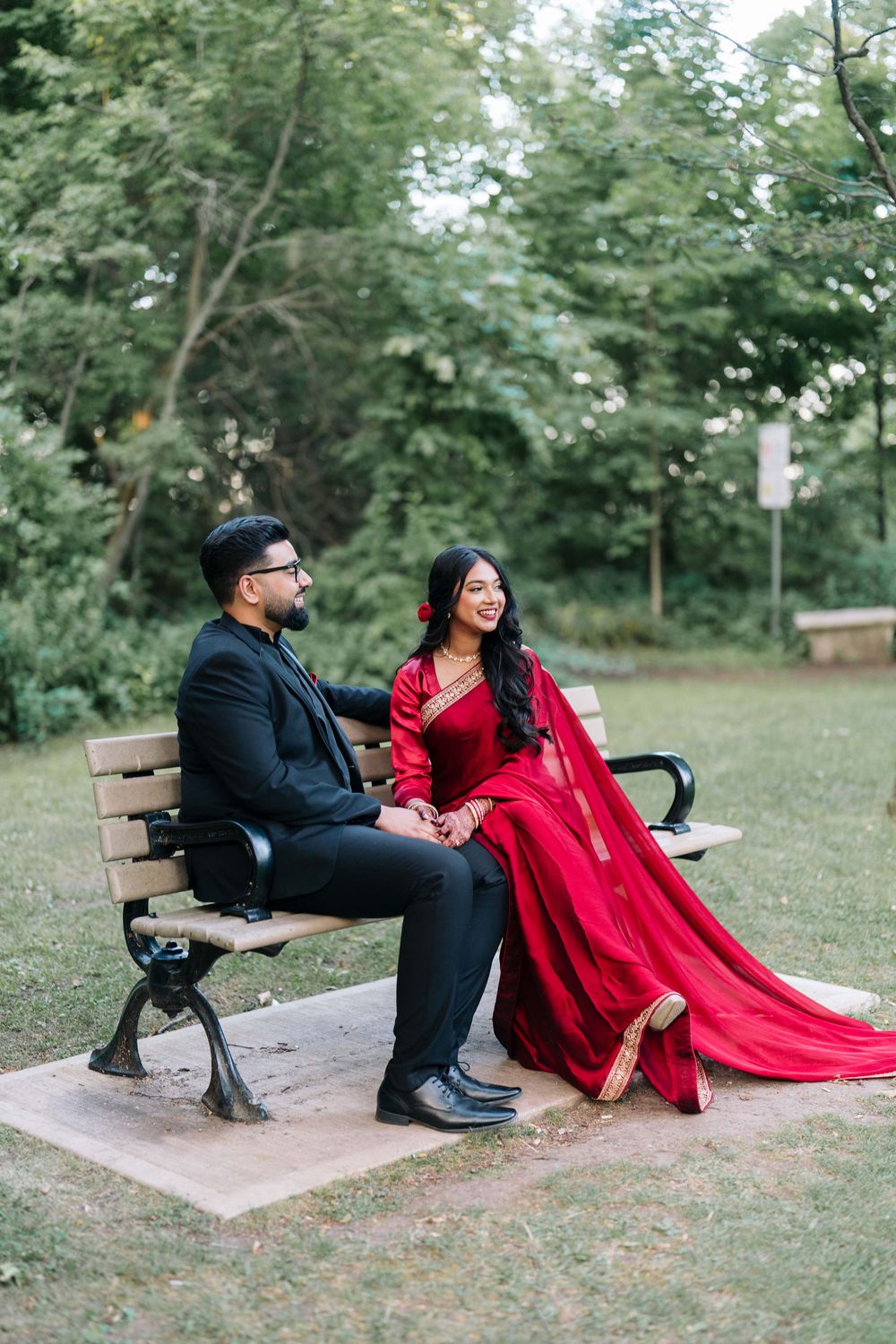 Couple in elegant attire sitting on a park bench, man in black suit and woman in red saree, surrounded by lush greenery.