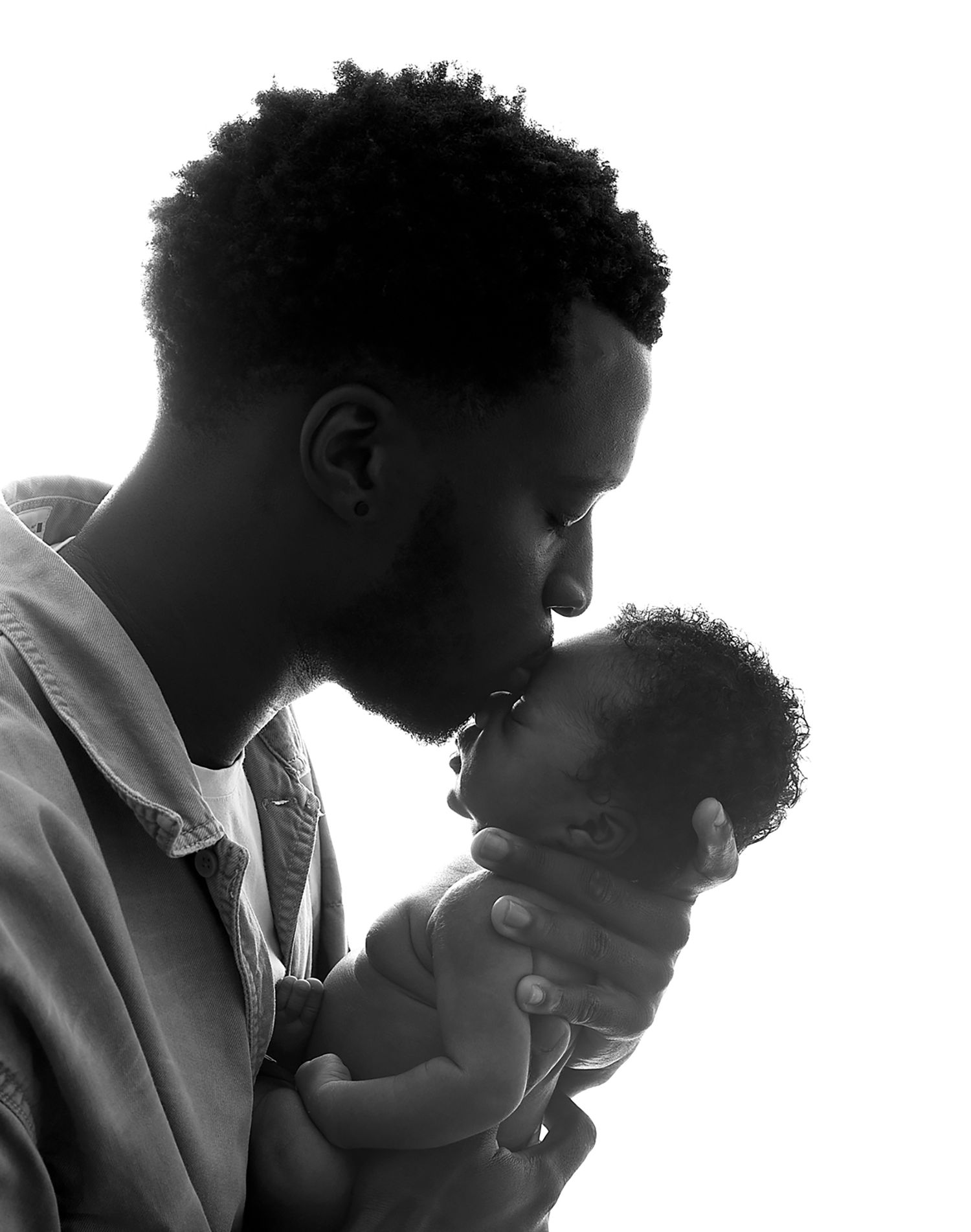 Black and white photo of a father tenderly kissing his newborn baby, captured in silhouette profile. During a newborn session in Bexley