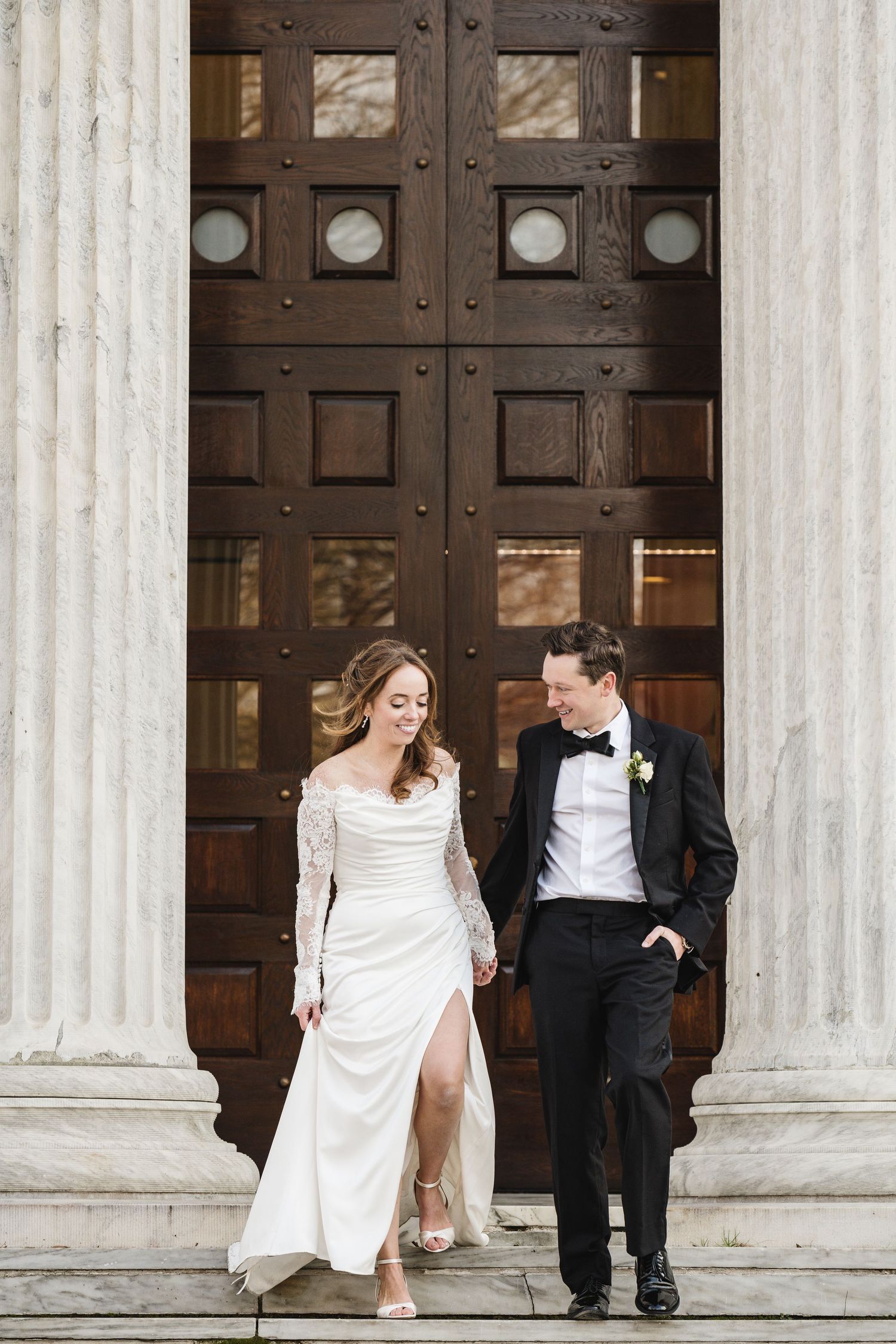 Bride in lace wedding gown and groom in tuxedo walking hand in hand before grand wooden doors and columns.