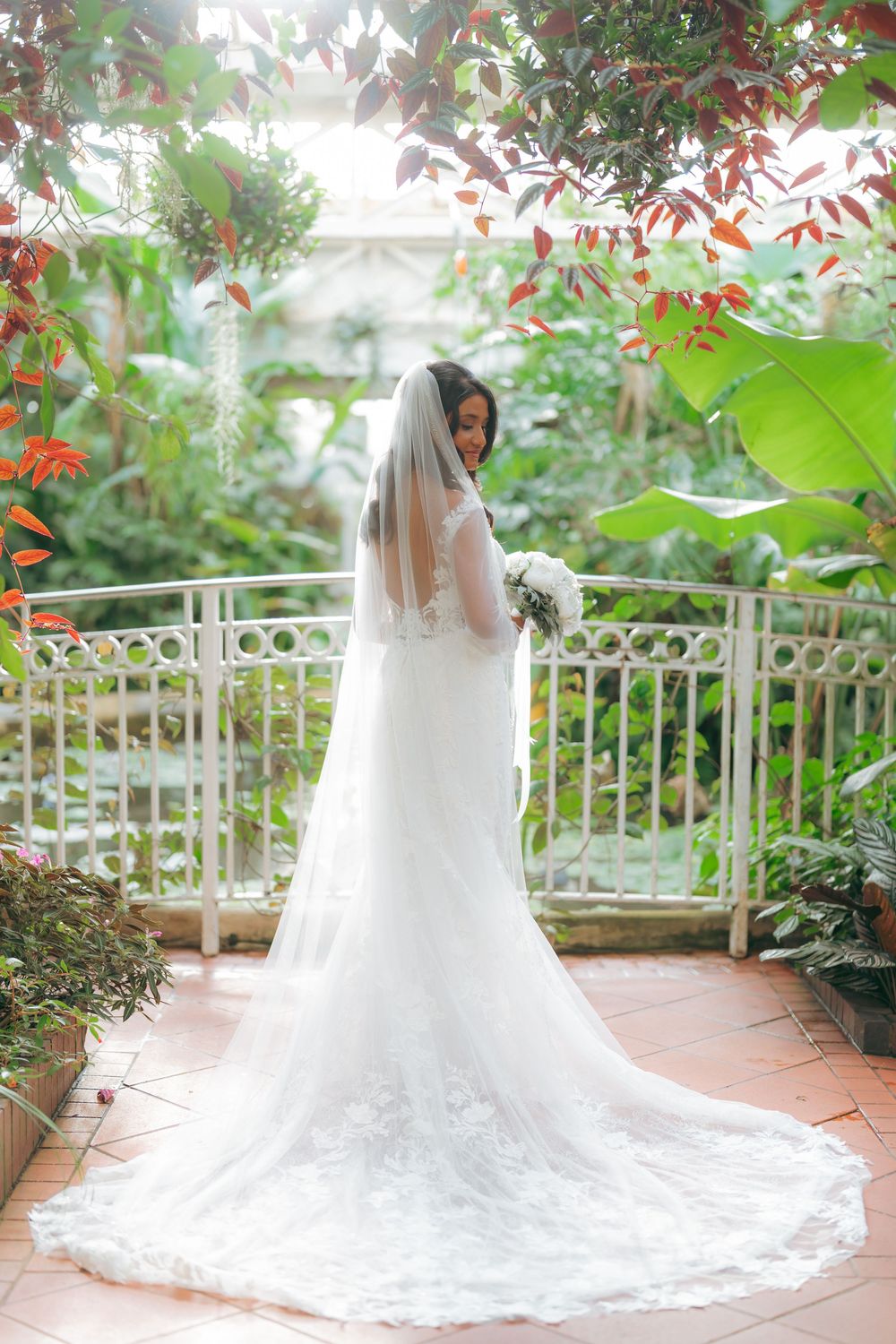 Bride in white lace gown and long veil holds bouquet on balcony surrounded by lush tropical greenery.