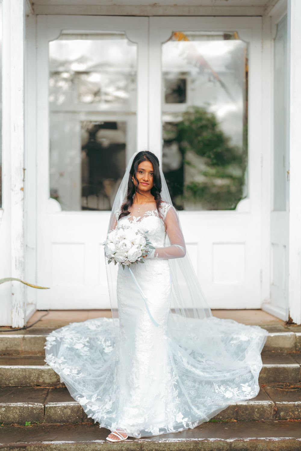 Bride in white lace mermaid gown with long veil holds bouquet on steps before white double doors.