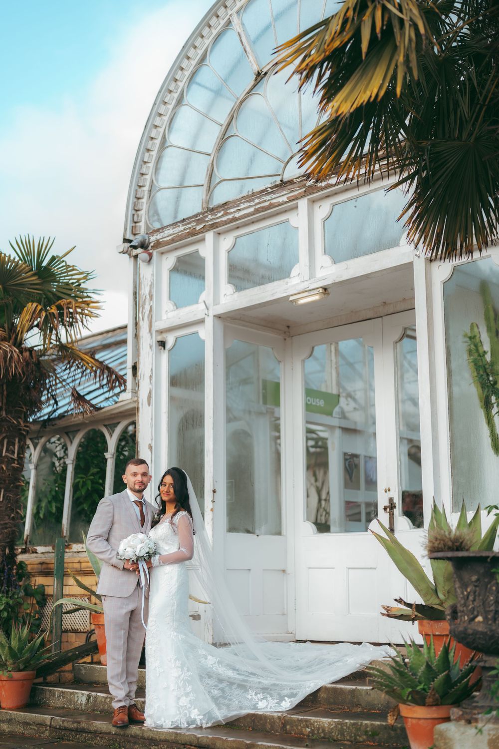 Bride and groom pose in front of a white Victorian greenhouse with palm trees on their wedding day.